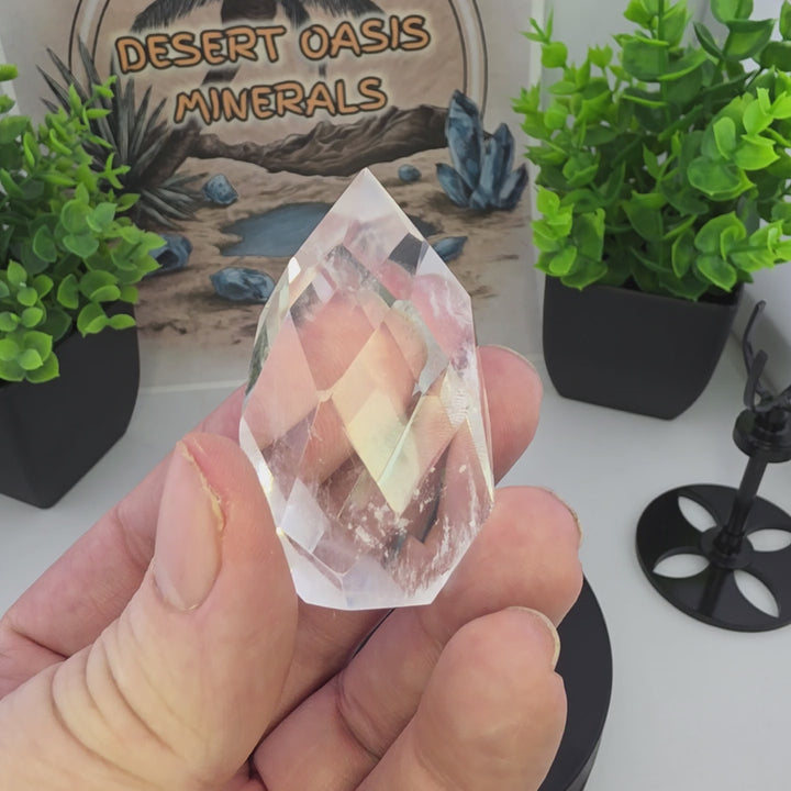Polished Faceted Clear Quartz Crystal Tear Drop with Stand held in hand with greenery and display elements in background.
