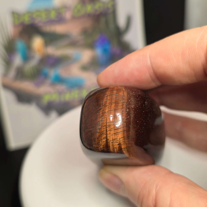 Polished Red Tiger's Eye freeform showcasing reddish-brown hues, held in hand against a desert-themed background.